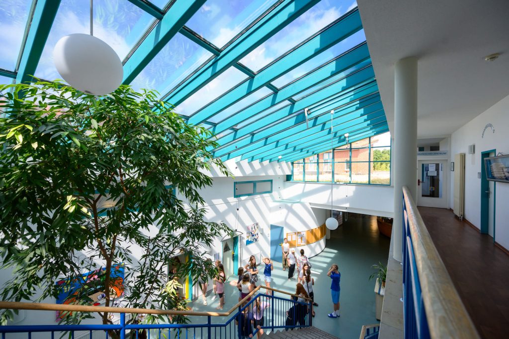 Moderner Innenbereich der Klinik: oben eine gläserne Dachschräge mit blauen Streben, durch die Sonnenlicht auf das Erdgeschoss fällt, eine Treppe führt nach unten, rechts ein Handlauf der ersten Etage mit Flur daneben.