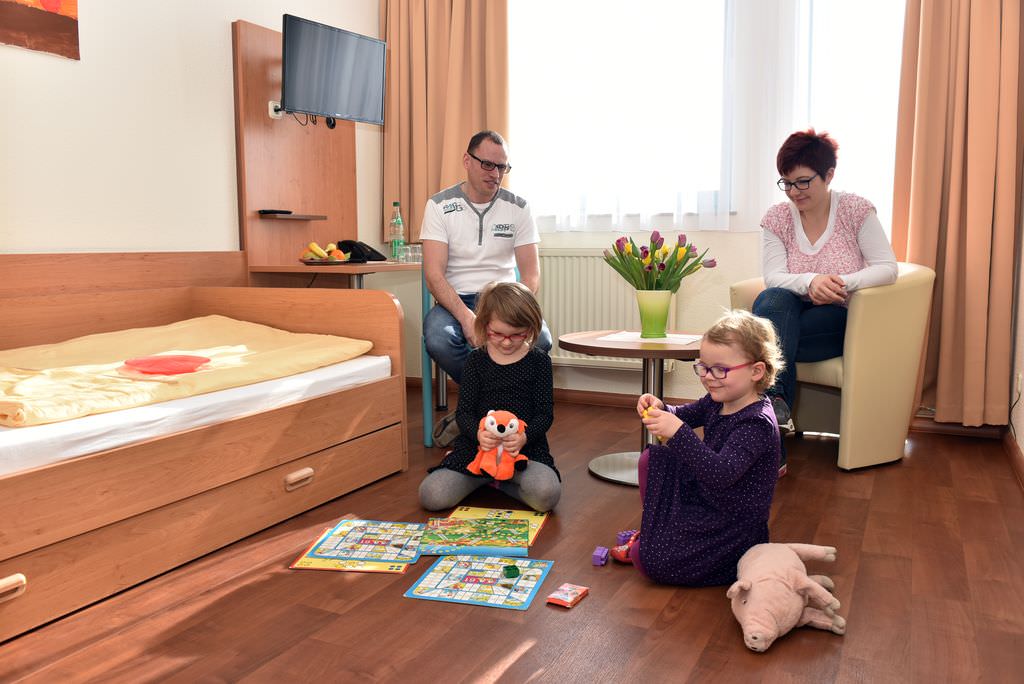 Zwei Kinder spielen auf dem Boden eines Appartements und werden dabei von Vater und Mutter beobachtet, die auf Sessel und Stuhl sitzen.