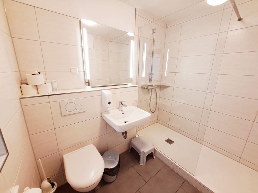 Badezimmer mit weißen Kacheln, Handwaschbecken in der Mitte, links eine Dusche, rechts eine Toilette.