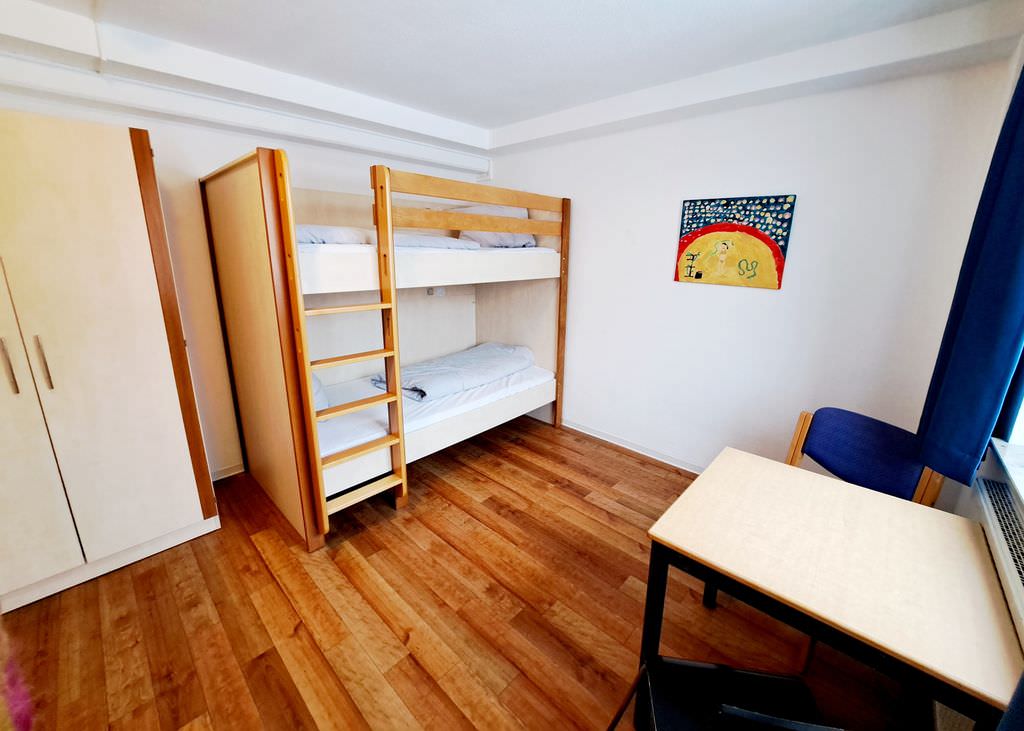Ein Etagenbett in der Mitte vor Kopf, links davon ein heller Schrank, rechts davon ein Kindergemälde einer Zirkusszene, rechts im Vordergrund ein Tisch und ein Stuhl.