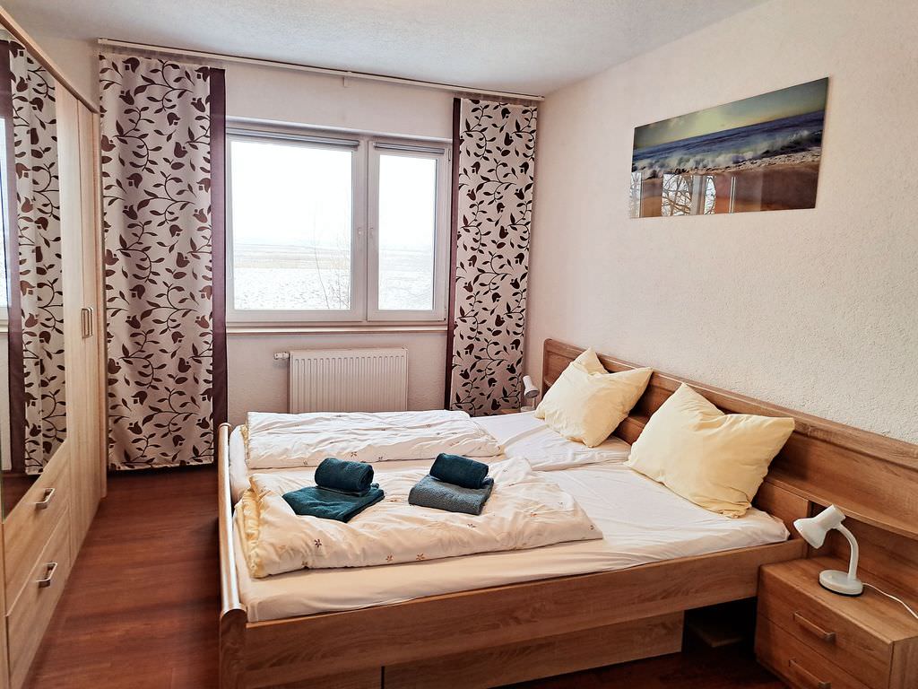 Ein Schlafzimmer mit moderner Einrichtung in Holz: ein Doppelbett, ein Kleiderschrank, ein Fenster mit floral gemusterten, geöffneten Vorhängen.