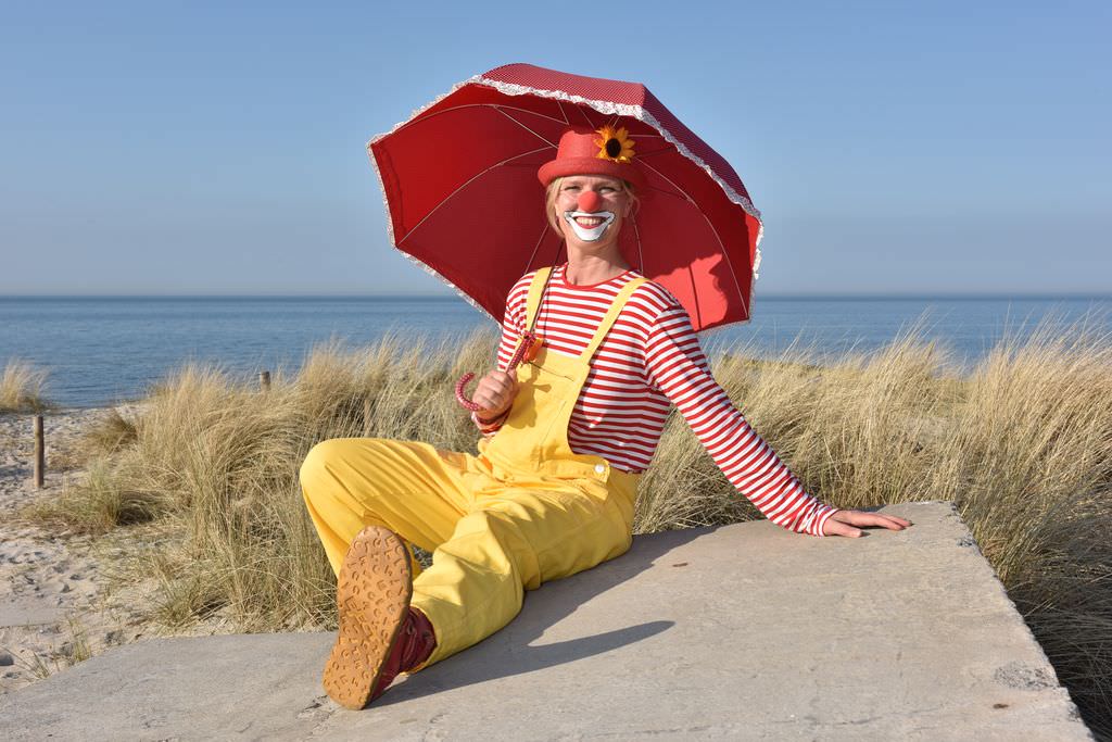 Clown mit gelber Latzhose, rot-weiß gestreiftem Shirt, rotem Hut mit Sonnenblume und rotem Sonnenschirm sitzend vor Gräsern am Ostseestrand.