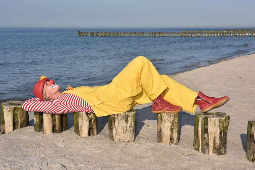 Clown mit gelber Latzhose, rot-weiß gestreiftem Shirt, rotem Hut mit Sonnenblume liegend auf niedrigen Holzstämmen am Ostseestrand. 