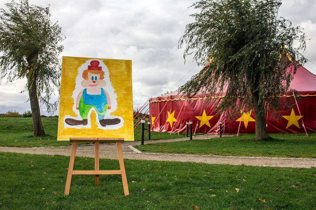 Ein gemalter Clown auf einer Staffelei vor einem roten Zirkuszelt mit gelben Sternen.