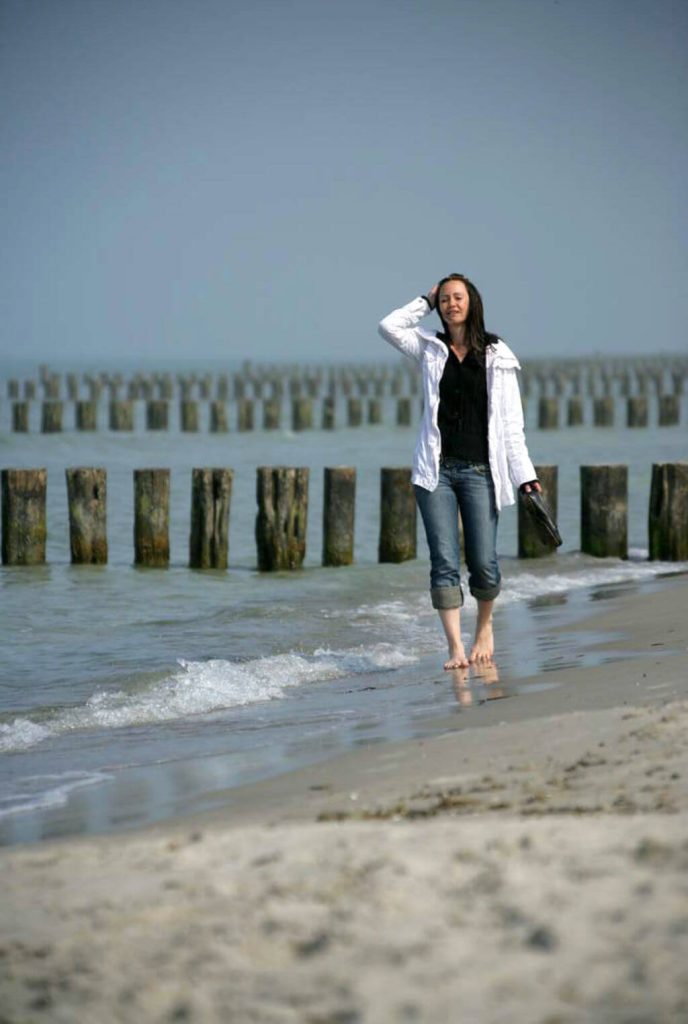 Eine Frau in weißer Jacke spaziert barfuß durchs Wasser der Ostsee am Strand.