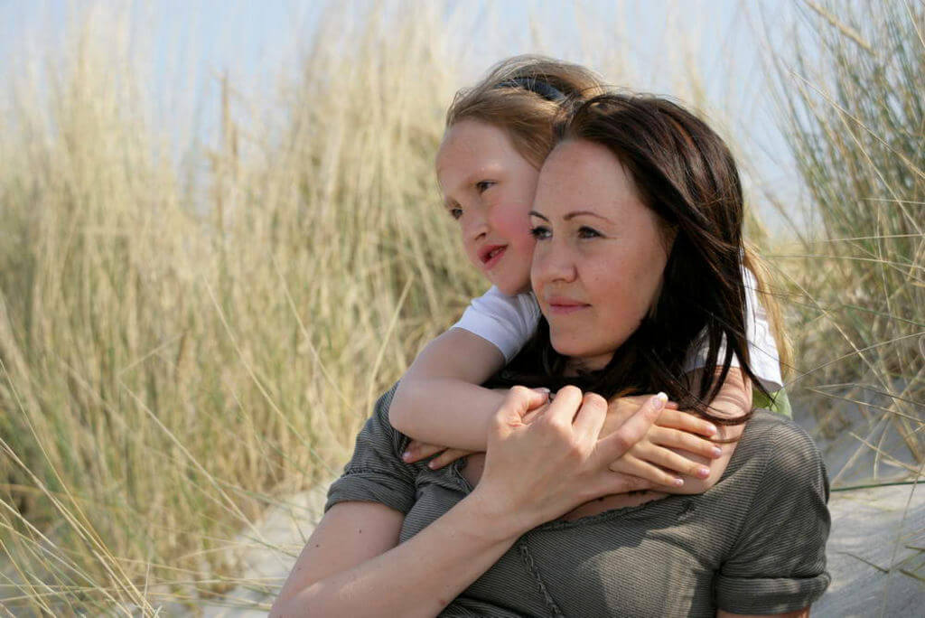 Eine Mutter sitzt im Dünengras und wird von hinten von ihrer Tochter umarmt.