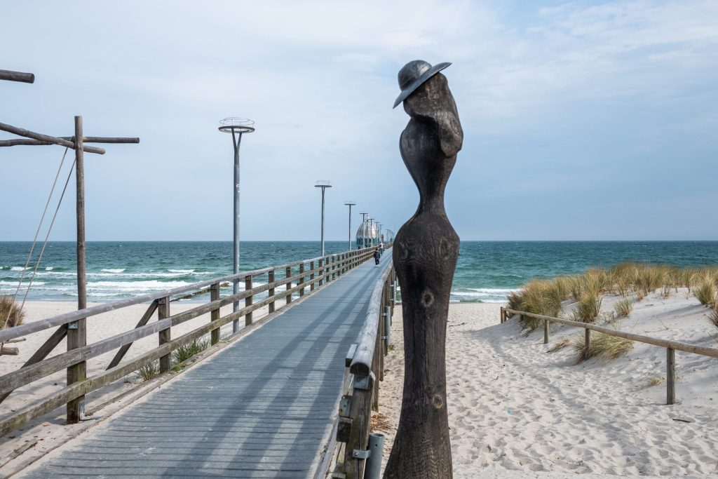 Ein Holzsteg führt über den hellen Sandstrand in die Ostsee bei Zingst. Rechts ist eine Holzskulptur zu erkennen.