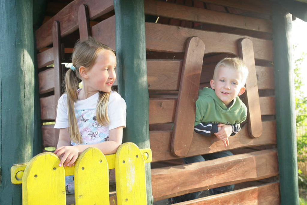 Ein Junge und ein Mädchen an einem Holzklettergerüst: Der Junge schaut durch ein Fenster hinaus, das Mädchen sitz hinter einem gelben Holzgartenzaun und schaut ihn an.