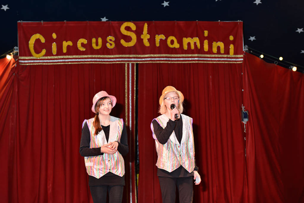 Zwei Frauen mit silber- und goldglänzenden Westen und Hüten vor einem roten Zirkusvorhang über dem Circus Stramini geschrieben steht.