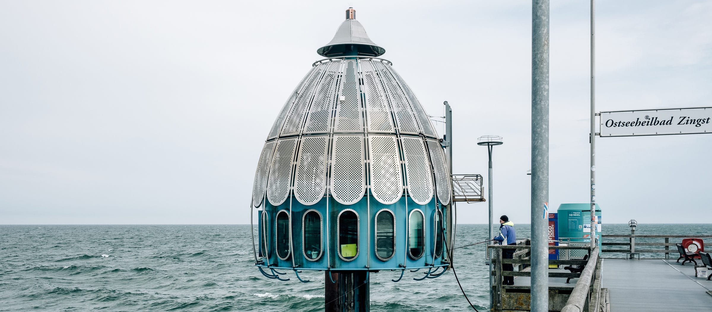 Die Tauchgondel Zingst: kuppelförmige Metallstruktur im Meer, zugänglich über einen Steg mit Personen.