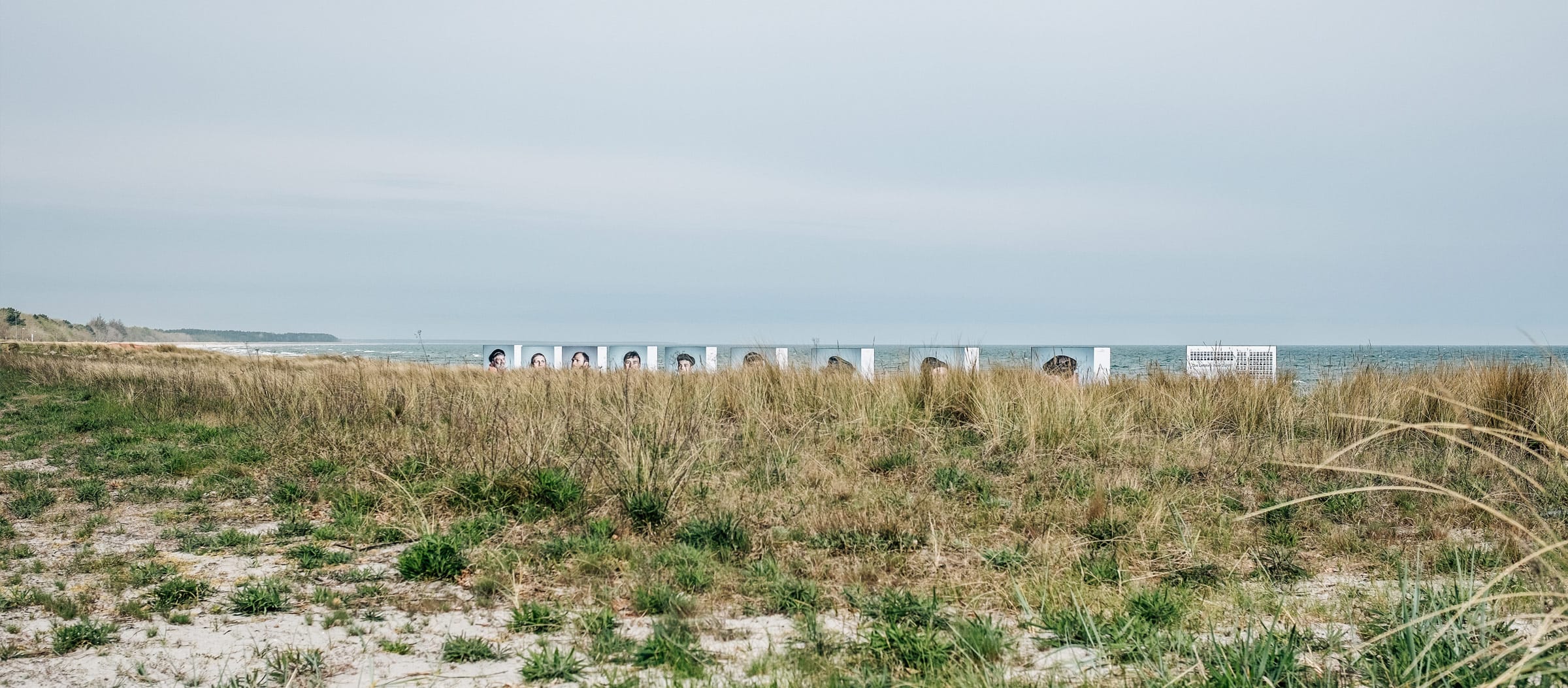 Strandlandschaft mit hohen Gräsern und einer Reihe von Kunstinstallationen/Portraits von Menschen an der Ostsee unter bewölktem Himmel.