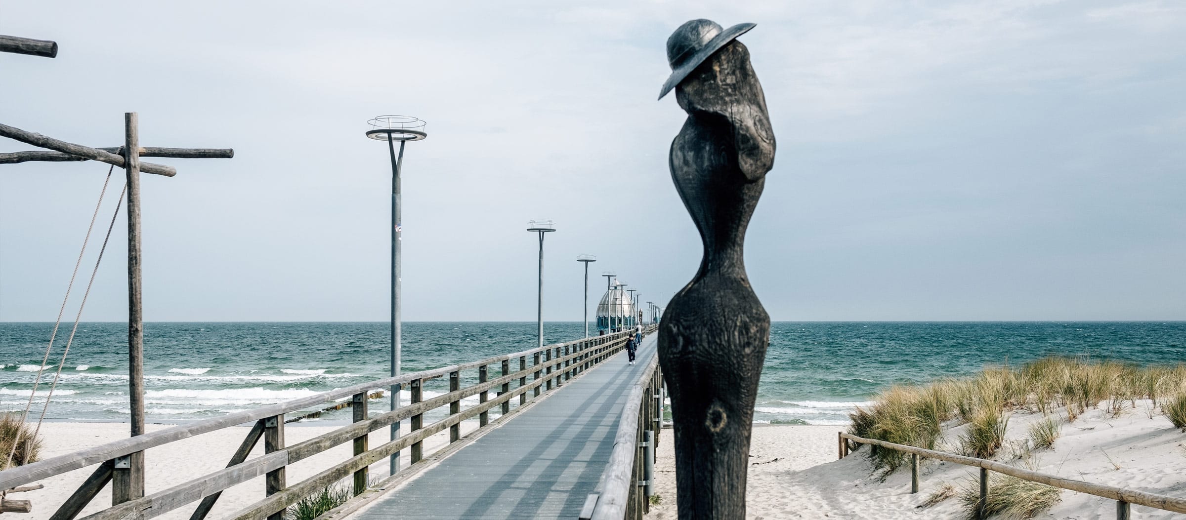 Die Seebrücke von Zingst: ein Steg der hinaus auf die Ostsee führt, rechts um Vordergrund eine Holzskulptur, die einen Menschen darstellt.