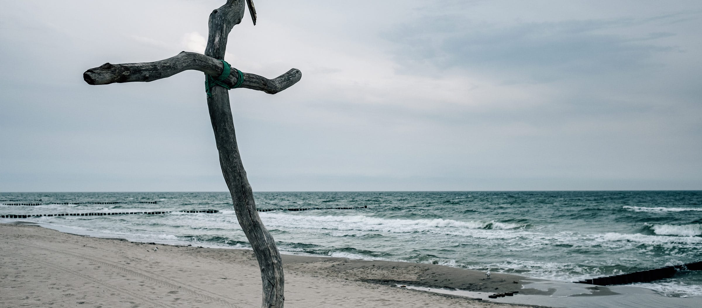 Ein krummes Holzkreuz am Strand an der leicht stürmischen Ostsee, die niedrige Wellen schlägt.