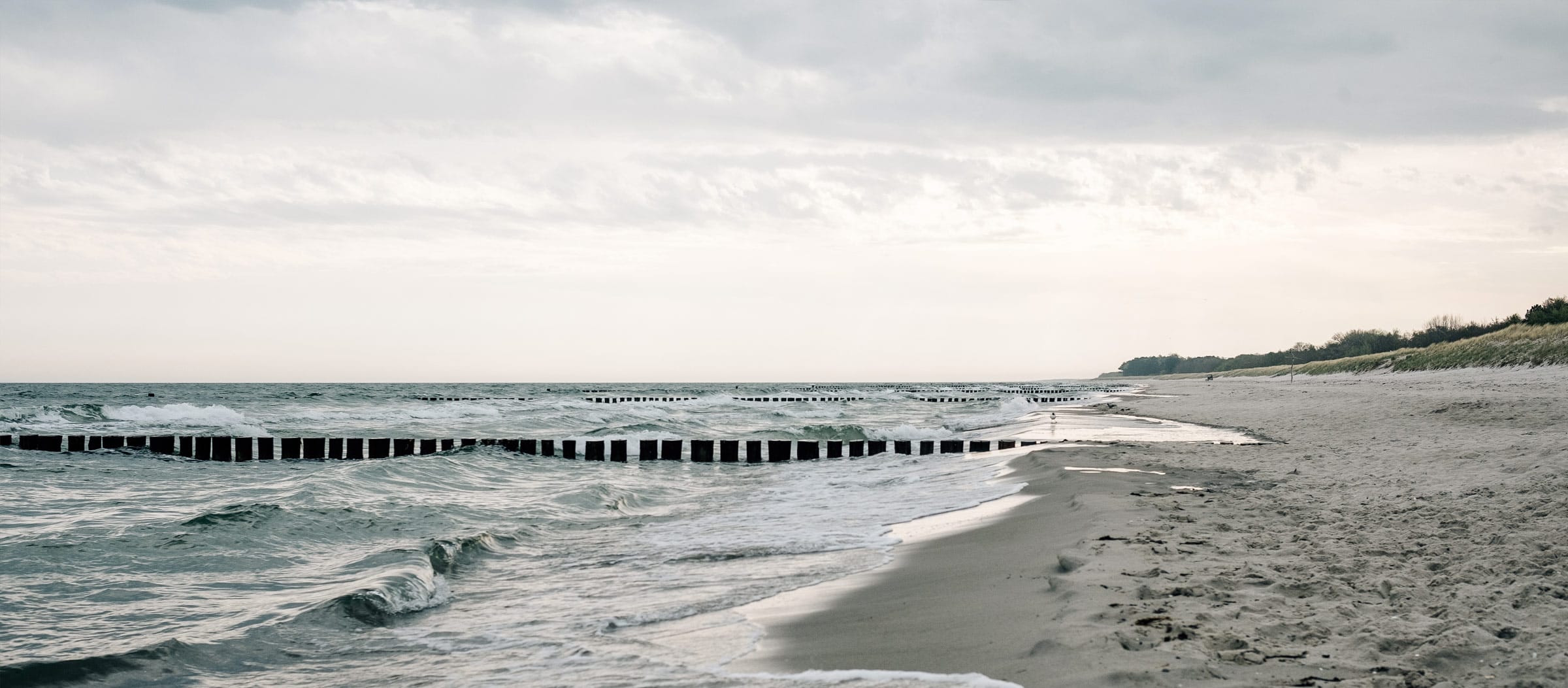 Links die etwas stürmische Ostsee, rechts ein Ostsee-Sandstrand. Über allem ein leicht bewölkter Himmel.