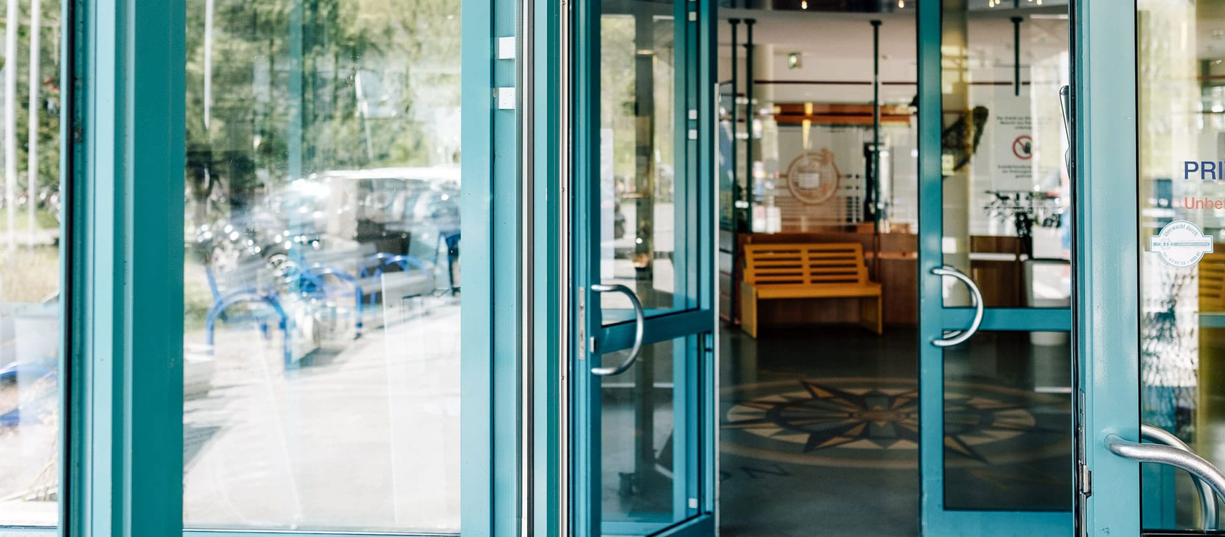 Gläserne, zur Hälfte geöffnete Flügel-Eingangstüre, Blick auf Holzbank im Gebäudeinneren.