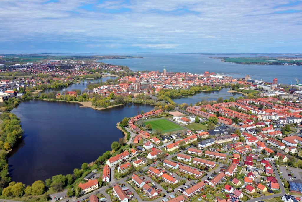Luftbildaufnahme von Stralsund: Wohngebiete und die historische Altstadt mit Kirchtürmen, umgeben von Wasser. Links ein großer See im Vordergrund, im Hintergrund ein großes Gewässer, der Strelasund, eine Verbindung zur Ostsee. Der Himmel ist blau mit Wolken.