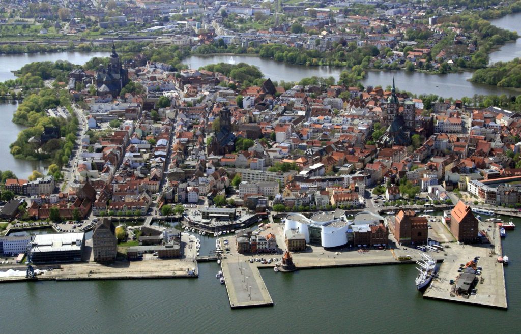 Luftbildaufnahme der historischen Altstadt von Stralsund, von Gewässern umgeben. Zu erkennen sind Bauwerke der Backsteingotik und Kirchtürme, unten der Hafen und die Marina von Stralsund.
