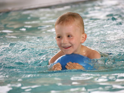 Ein lachender Junge mit einem blauen Ball schwimmend im Schwimmbecken.