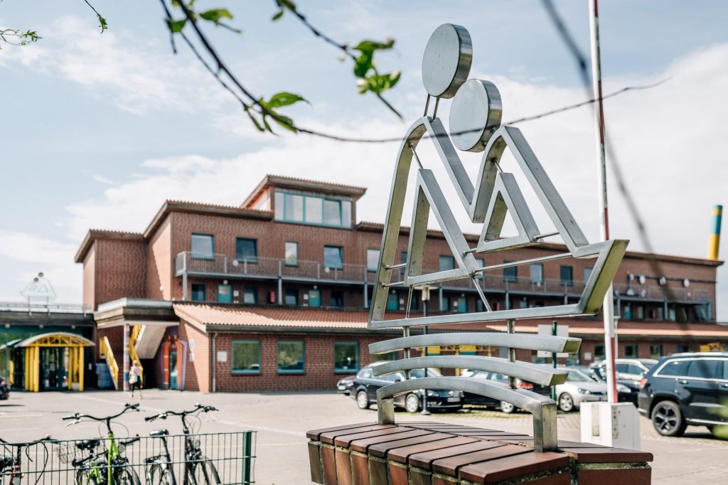 Hauptgebäude der Ostseeklinik Zingst: ein modernes Backsteingebäude, davor ein Parkplatz mit Autos, ganz im Vordergrund eine silberne Metallskulptur, die zwei einander zugewandte Menschen symbolisiert.