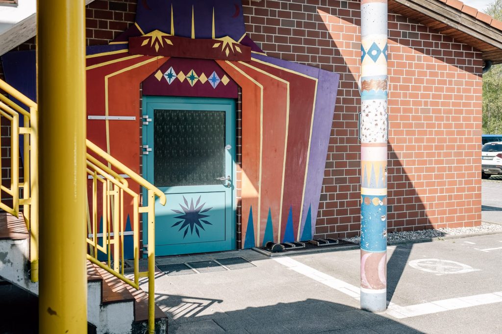 Kindgerecht und farbenfroh gestalteter Seiteneingang der Ostseeklinik Zingst: eine blaue Tür mit Sternmotiv ist von einer bühnenartigen, mehrfarbigen Holzverkleidung in Rot-, Orange- und Lilatönen mit Rautenmustern umgeben. 