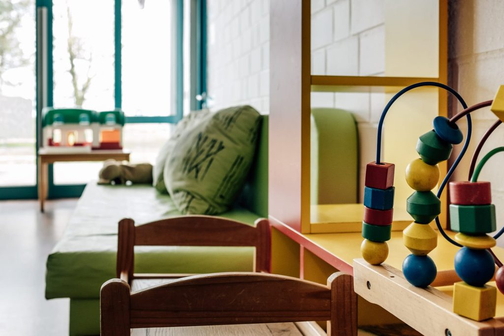 Aufenthaltsraum mit gepolsterter Sitzbank und Kissen rechts, davor ein niedriger Holztisch mit Kinderstühlen, rechts im Vordergrund ein bunter Kinder-Holzspielzeug.