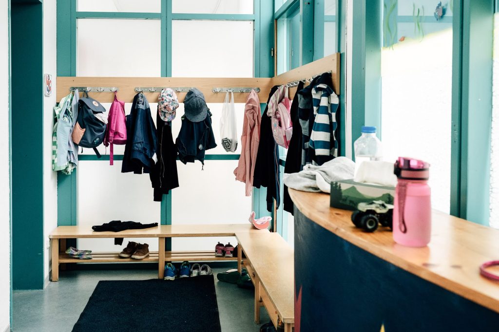 Eine Holzgarderobe über Eck vor halbtransparenten, bodentiefen Fenstern. An der Garderobe hängen verschiedene Kinder-Kleidungsstücke.