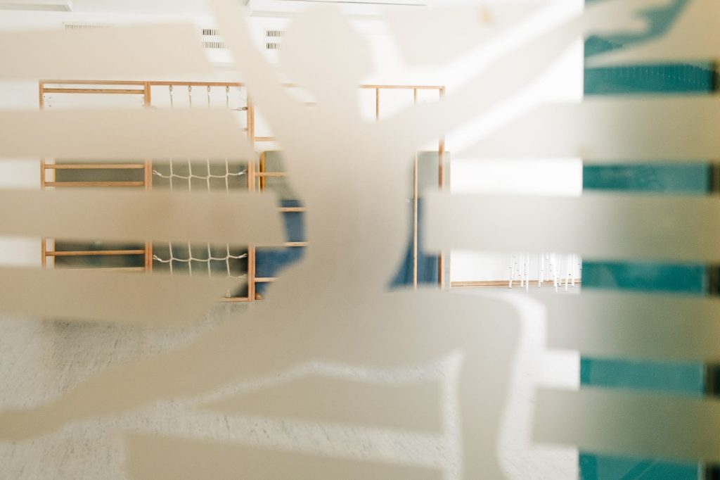 Eine Glasfläche mit einem stilisierten, turnenden Menschen darauf. Durch das Glas ist ein Sport- und Gymnastikraum mit Sprossenwand zu sehen.