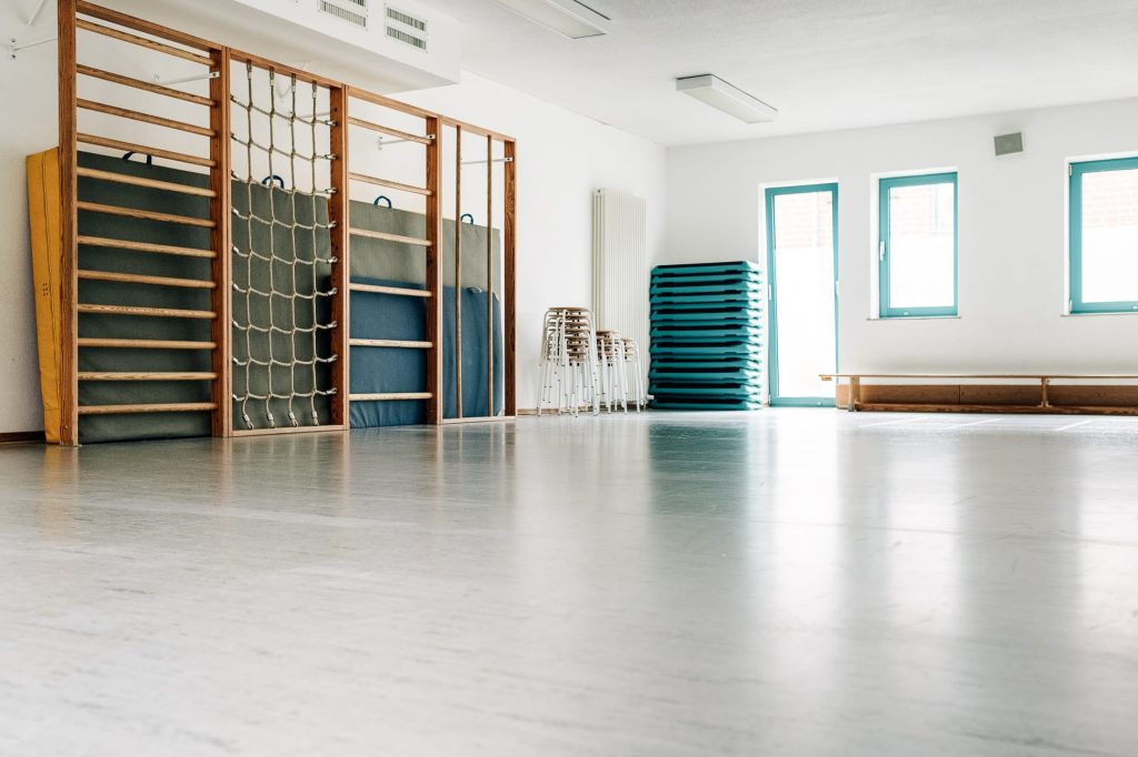Die Sporthalle der Ostseeklinik: links einer Sprossenwand samt Kletterseilnetz, im Hintergrund vor Kopf gestapelte Hocker und Turnmatten sowie eine Holzbank.