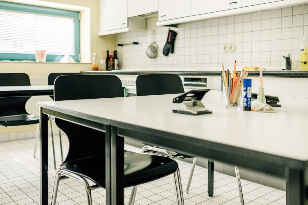 Lehrküche der Ostseeklinik mit langer Küchenzeile rechts im Hintergrund, im Vordergrund ein Tisch, auf dem sich Schreibutensilien und ein Locher befinden, vor dem Tisch zwei Stühle.