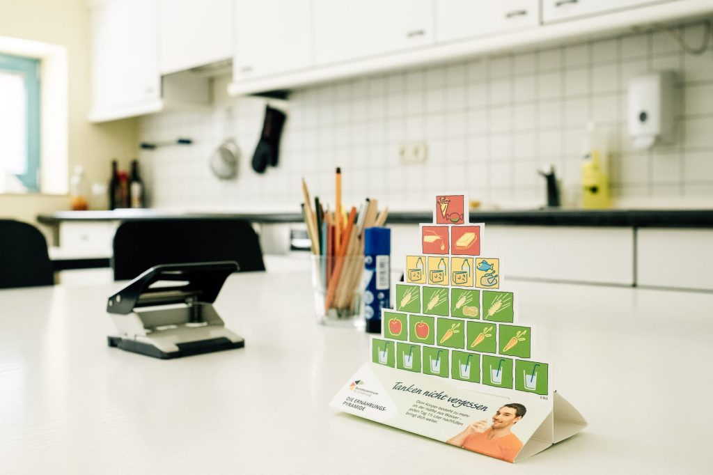 Eine Küche mit großer Küchenzeile im Hintergrund, im Vordergrund ein Tisch mit einer Pyramide aus Memory-Karten, auf denen sich unterschiedliche Gemüsesorten abgebildet sind sowie ein Glas mit Stiften und ein Locher.