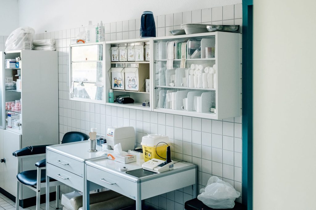 Ein medizinisches Behandlungs- und Untersuchungszimmer mit weißer Einrichtung: Wandregale, Regalschrank und Tisch mit medizinischen Utensilien.