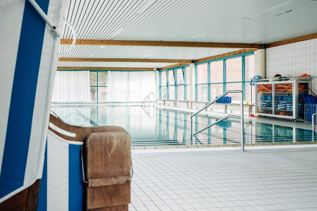 Das helle Schwimm- und Bewegungsbad mit bodentiefen Fenstern ringsum, mit Geländern, die ins Schwimmbecken führen und mit Trainingsgeräten rechts in einem Gitterschrank.