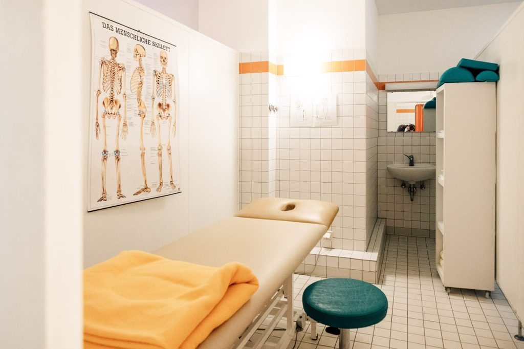 Ein Behandlungs- und Untersuchungszimmer: links eine Liege mit einem Hocker davor, an der Wand dort ein Schaubild mit einem menschlichen Skelett, hinten vor Kopf ein Waschbecken.