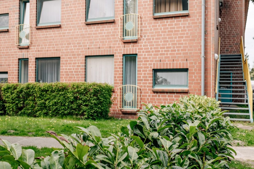 Anschnitt einer Backsteinfassade mit Fenstern und kleinen Balkonen, rechts davon eine Treppe, davor zwei Bäume.