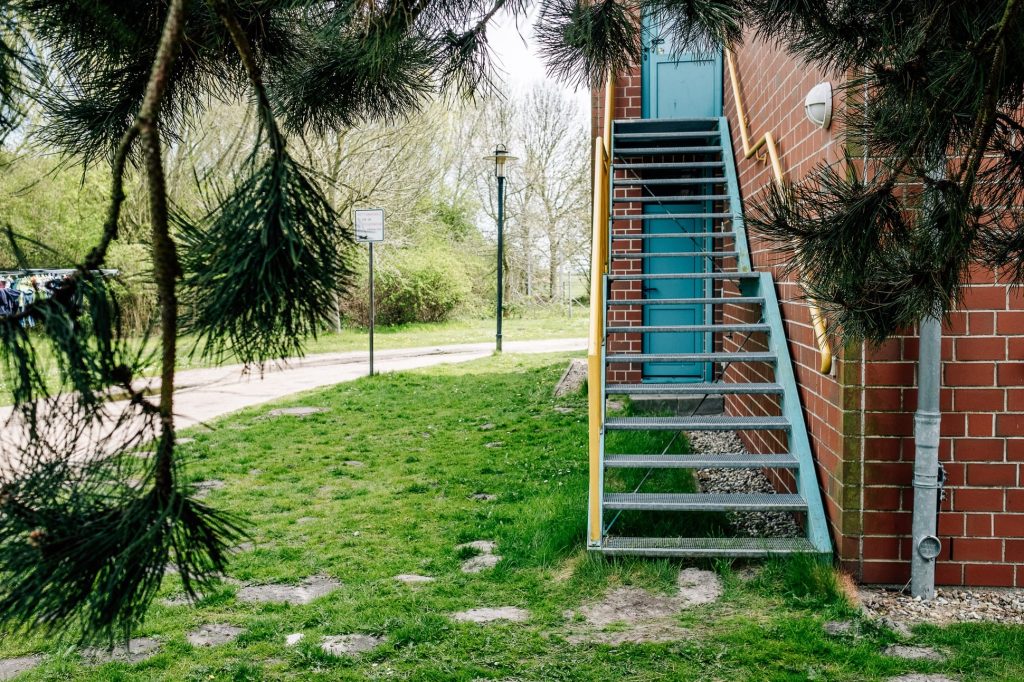 Rechts eine Treppe entlang einer Backsteinfassade, die zu einer Türe führt. Links eine Wiese mit Fußweg und Bäume.