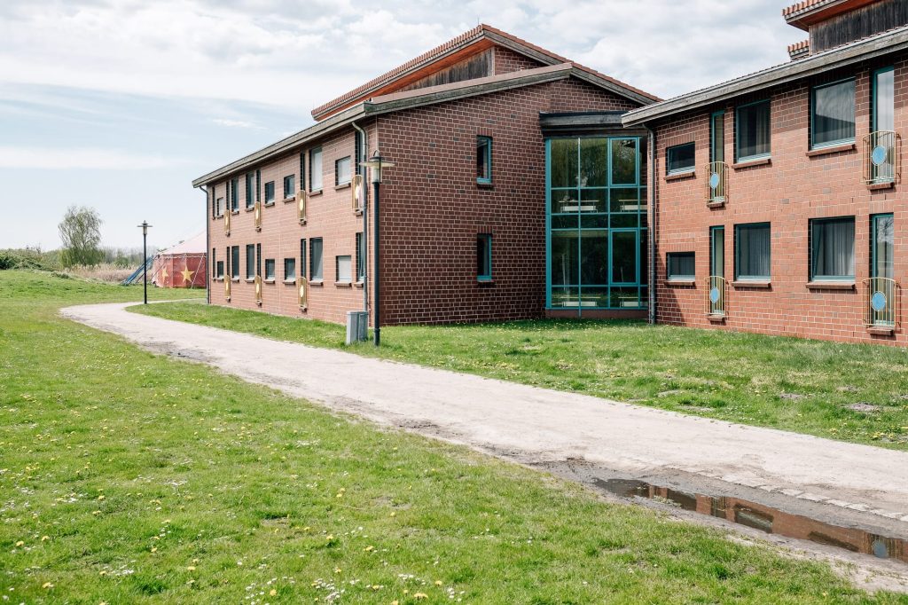 Zwei zweistöckige Backstein-Wohngebäude der Ostseeklinik Zingst rechts, links davon eine Wiese, durch die ein Weg führt. im Hintergrund ein rotes Zirkuszelt.