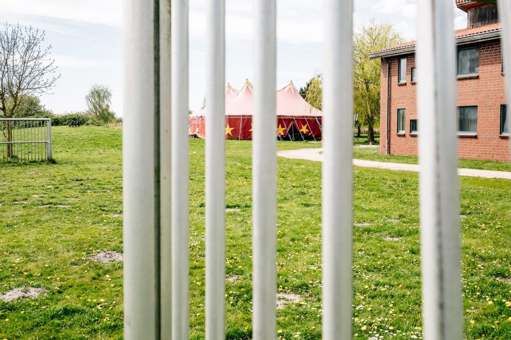 Blick durch Metallstäbe hindurch auf eine grüne Wiese mit rotem Zirkuszelt in der Mitte, rechts davon ein Backsteinhaus im Anschnitt.
