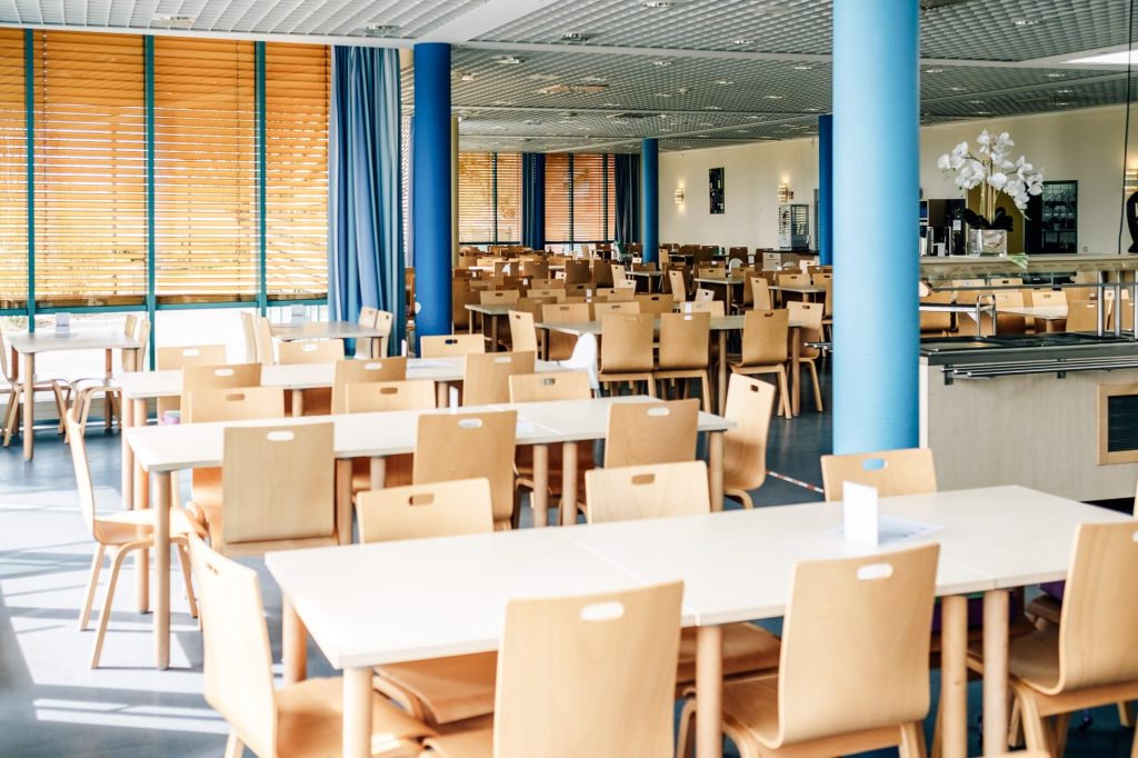 Der helle Speisesaal der Ostseeklinik Zingst mit weißen Tischen und hellen Holzstühlen, Fensterjalousien dämpfen das einfallende Sonnenlicht.