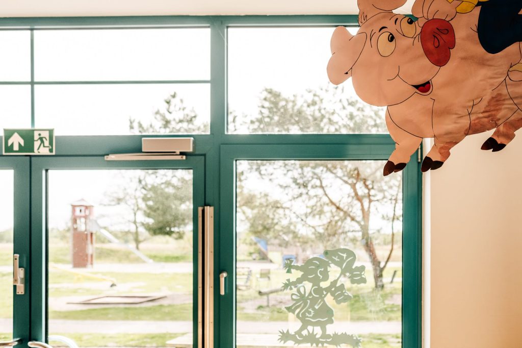 Blick durch eine gläserne Türe auf den Kinderspielplatz der Klinik, im Vordergrund scheint ein Glücksschwein aus Papier zu schweben.