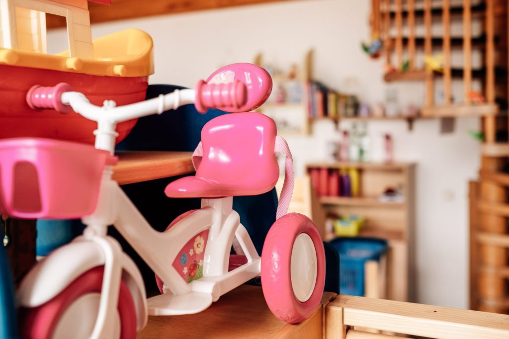 Kinderspielzeug: ein rosafarbenes, kleines Plastikdreirad, links daneben ein kleines Schiff im Anschnitt, im Hintergrund Regale.