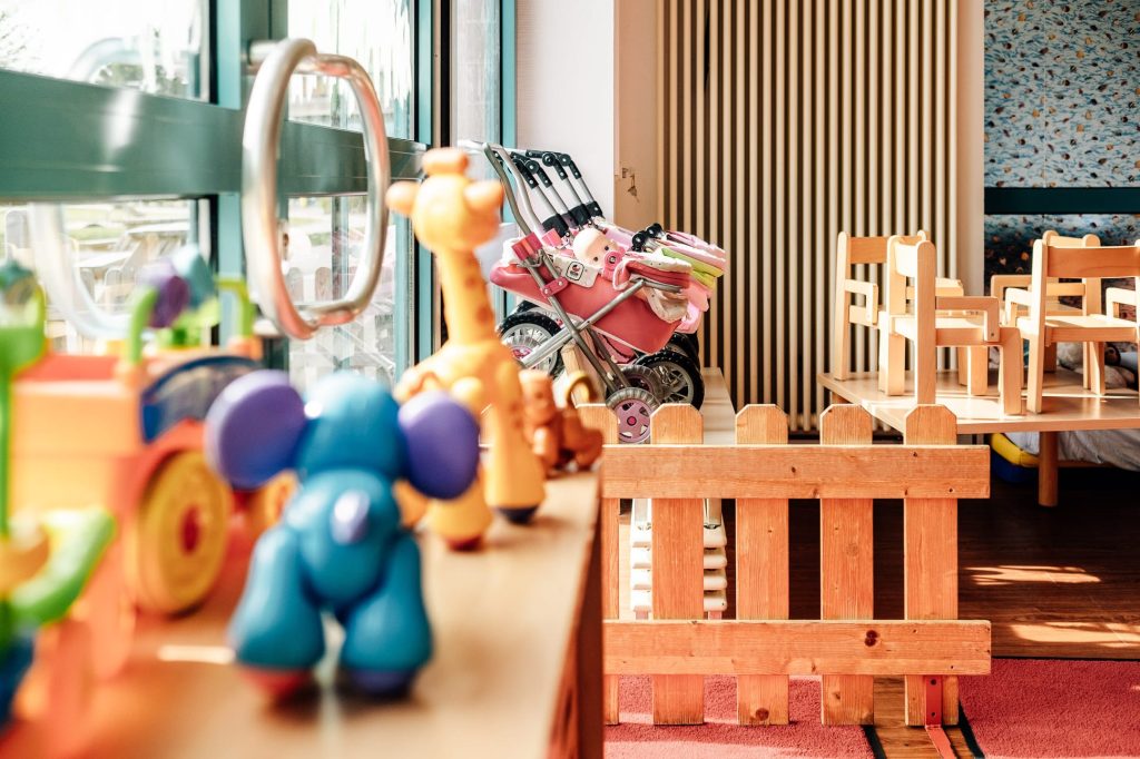 Links Spielfiguren für Kinder, dahinter Kinderwagen, rechts ein niedriger Holztisch mit kleinen Kinderstühlen darauf.