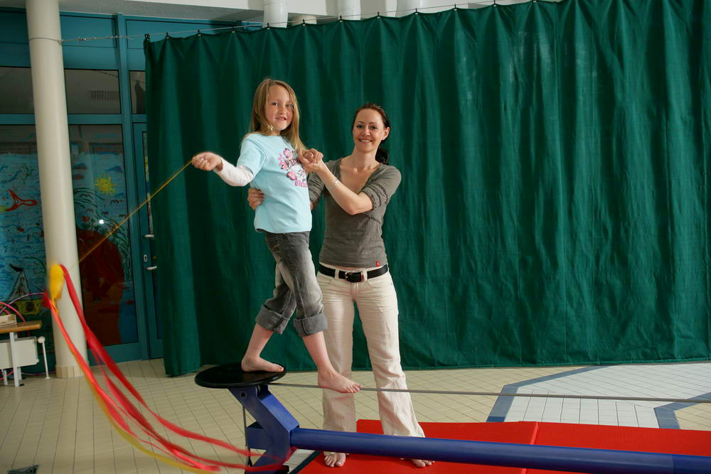 Ein Mädchen balanciert auf einem Seilt und wir dabei von einer Frau gehalten und unterstützt.