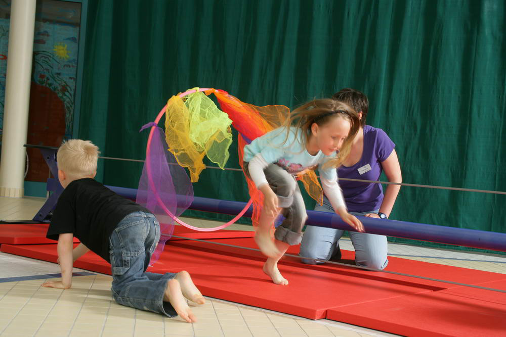 Ein Mädchen springt durch einen Zirkusreifen, der von einer Frau gehalten wird, davor krabbelt ein Junge auf allen Vieren.