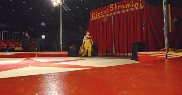 Ein Clown mit Koffer betritt die Manege, rechts davon ein roter Vorhang, darüber der Schriftzug Circus Stramini