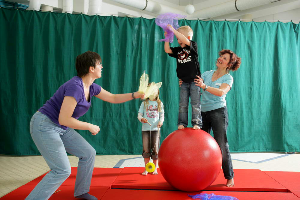 Ein Junge steht auf einem roten Gymnastikball und wirft ein Tuch. Er wird gehalten von einer Frau. Eine andere Frau versucht das Tusch zu fangen.